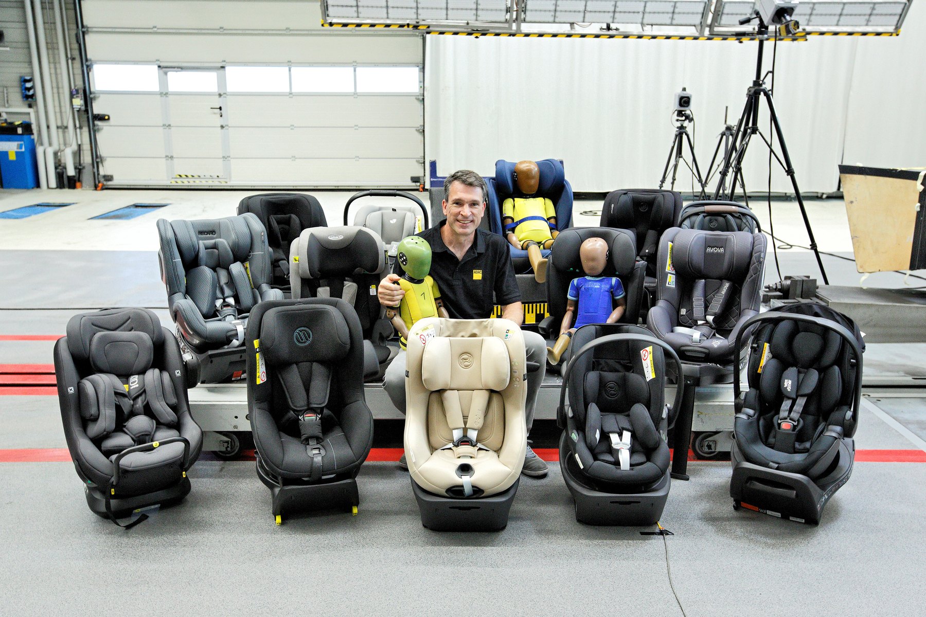 Les sièges auto du test d'automne 2026 alignés dans le hall d'essai de l'ADAC, avec trois mannequins de crash-test et un membre de l'équipe ADAC